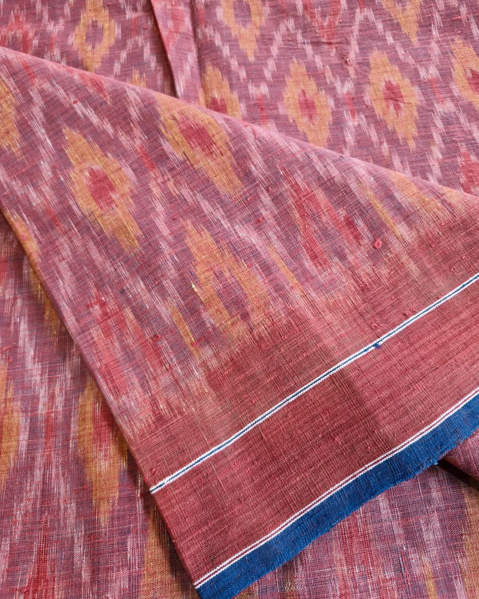 A close-up of a folded woven textile with a rust-red and orange ikat pattern, featuring a blue and white striped border.