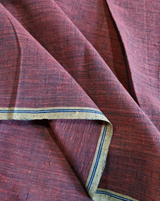 A close-up of draped textured fabric in a muted burgundy-red tone, with a folded section showing a narrow border stripe in blue and beige.