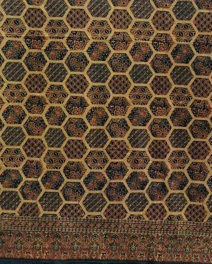 A close-up of a patterned textile or rug showing a repeating honeycomb-like geometric design in dark brown, black, beige, and muted red tones, with a decorative border along the bottom edge.