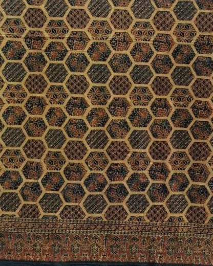 A close-up of a patterned textile or rug showing a repeating honeycomb-like geometric design in dark brown, black, beige, and muted red tones, with a decorative border along the bottom edge.