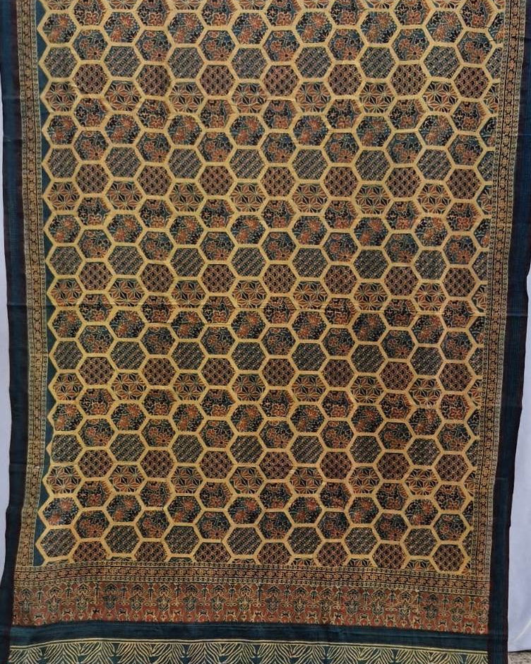 A traditional patterned textile, likely a saree or fabric panel, featuring a dense geometric honeycomb motif in black, brown, and gold, with a decorative border at the bottom.