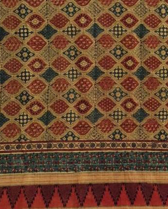 A close-up of a patterned textile or rug showing a repeating diamond motif in red, dark blue, beige, and brown, with a decorative border along the bottom.