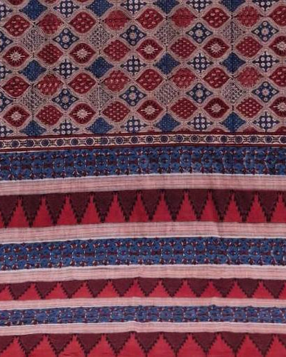 A close-up of a patterned textile or fabric with geometric motifs in red, blue, beige, and dark brown, including diamond, cross, and triangular stripe patterns.