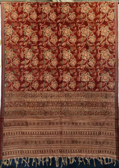 A traditional patterned textile, likely a saree or scarf, featuring a rich rust-red background with intricate floral motifs in beige, brown, and muted green. The bottom portion is adorned with multiple decorative borders in earthy tones and finished with a fringed edge.