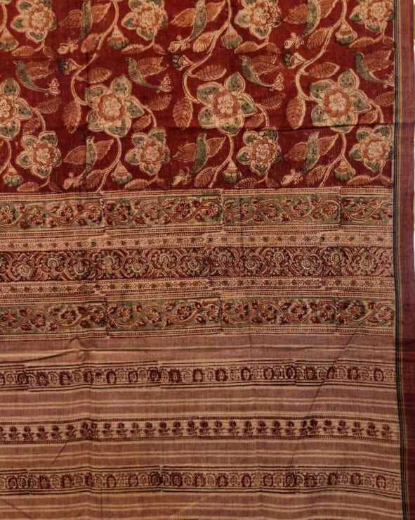 A close-up of a traditional patterned textile, likely a sari, featuring a deep red background with intricate floral motifs in beige, brown, and green, and a wide decorative border with tassels at the bottom.
