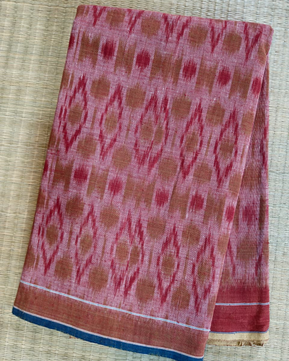 A folded piece of pink and rust-colored patterned fabric, likely a cotton saree or scarf, featuring a traditional ikat design and a blue border, laid flat on a woven straw mat.