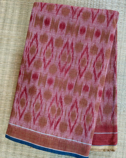 A folded piece of pink and rust-colored patterned fabric, likely a cotton saree or scarf, featuring a traditional ikat design and a blue border, laid flat on a woven straw mat.