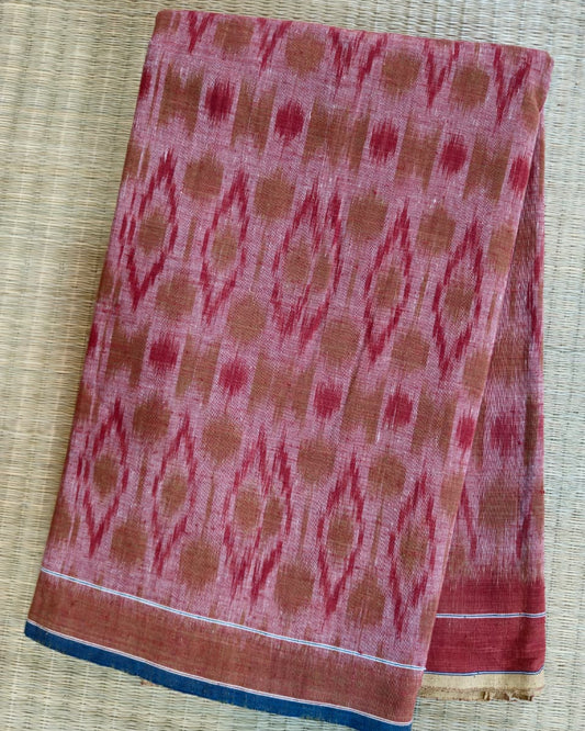 A folded piece of pink and rust-colored patterned fabric, likely a cotton saree or scarf, featuring a traditional ikat design and a blue border, laid flat on a woven straw mat.