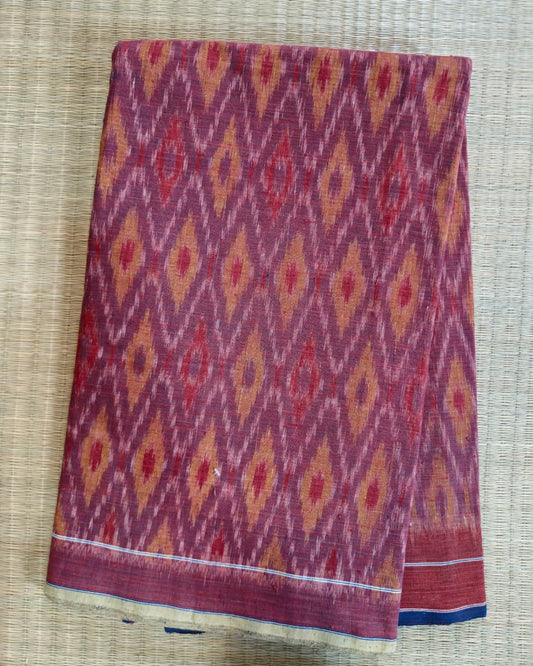 A folded piece of patterned fabric with an ikat-style diamond motif in burgundy, red, orange, and cream, displayed on a woven straw mat.