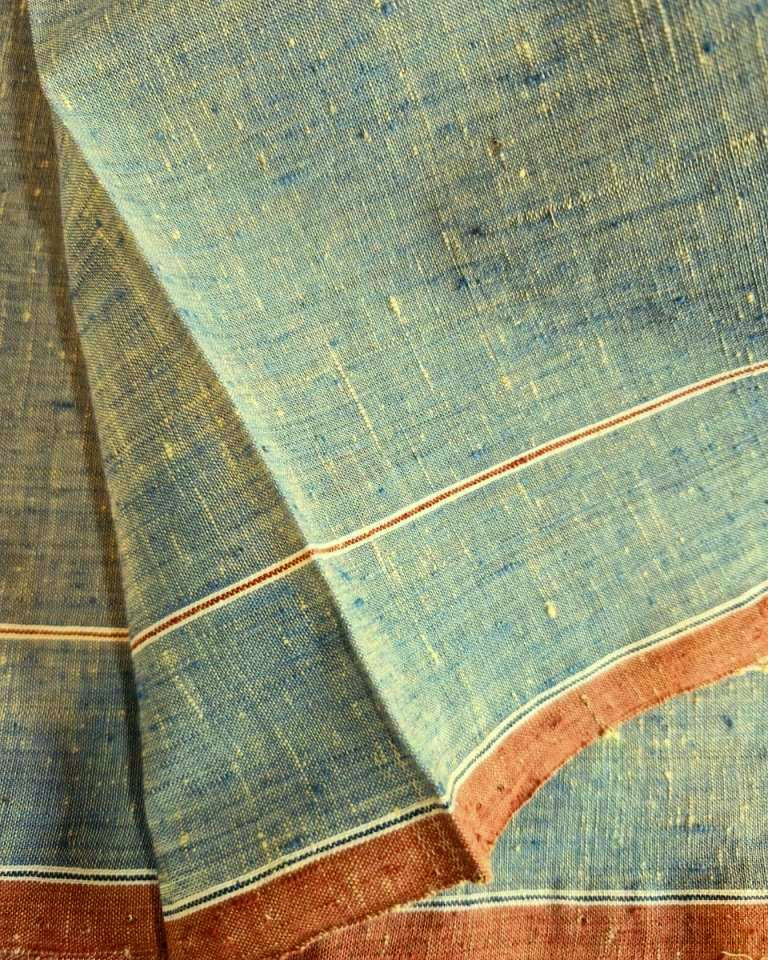 A close-up of a textured blue fabric with fine slub yarns, featuring a rust-red border with thin cream stripes.