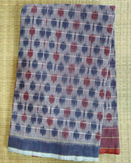A folded patterned scarf or shawl laid flat on a woven straw mat. The fabric has a muted blue-gray base with repeating red motifs and thin white grid lines, plus a darker blue border with small gold accents.