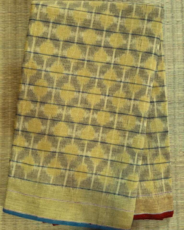 A folded yellow and brown patterned fabric, likely a saree or scarf, featuring a geometric ikat design with a blue and red border, laid flat on a woven straw mat.