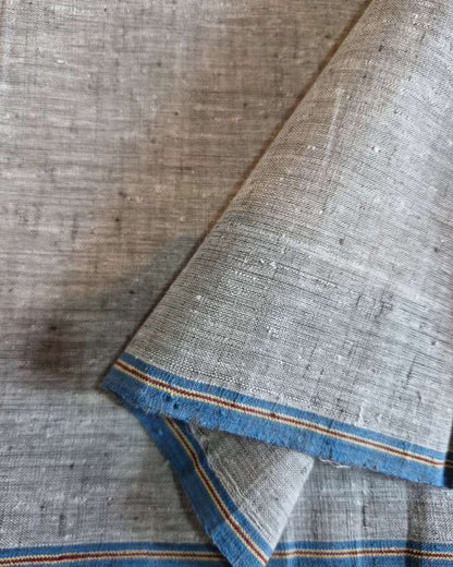 A close-up of a textured woven fabric, likely cotton or linen, in a light gray-beige tone with a contrasting blue border accented by thin orange stripes. The fabric is draped diagonally, showing its weave and texture.