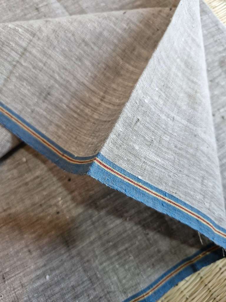 A close-up of a folded woven fabric with a light beige body and blue borders accented by thin orange and yellow stripes, laid on a rustic wooden surface.