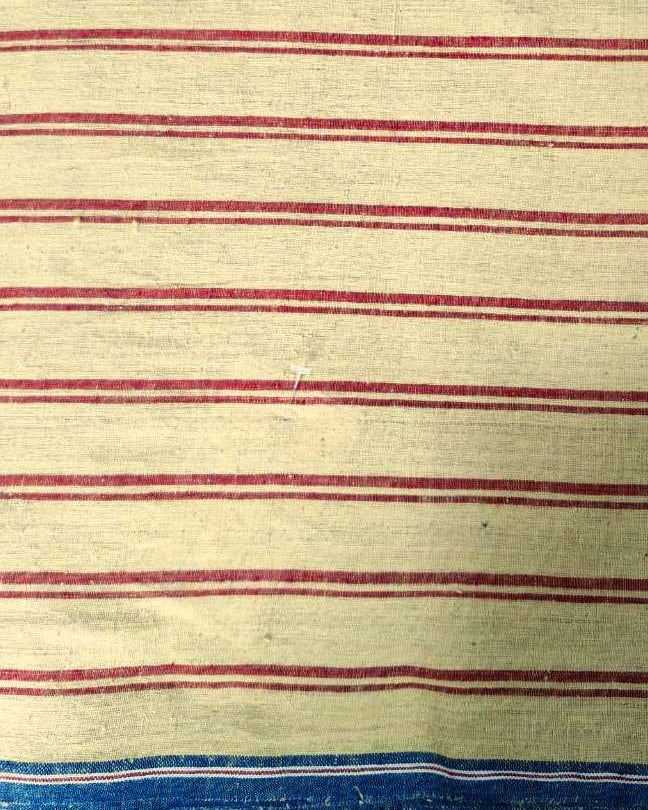 A close-up of a striped textile, likely a vintage striped blanket or towel, featuring horizontal red stripes on a cream-colored background, with a blue and white striped border at the bottom.