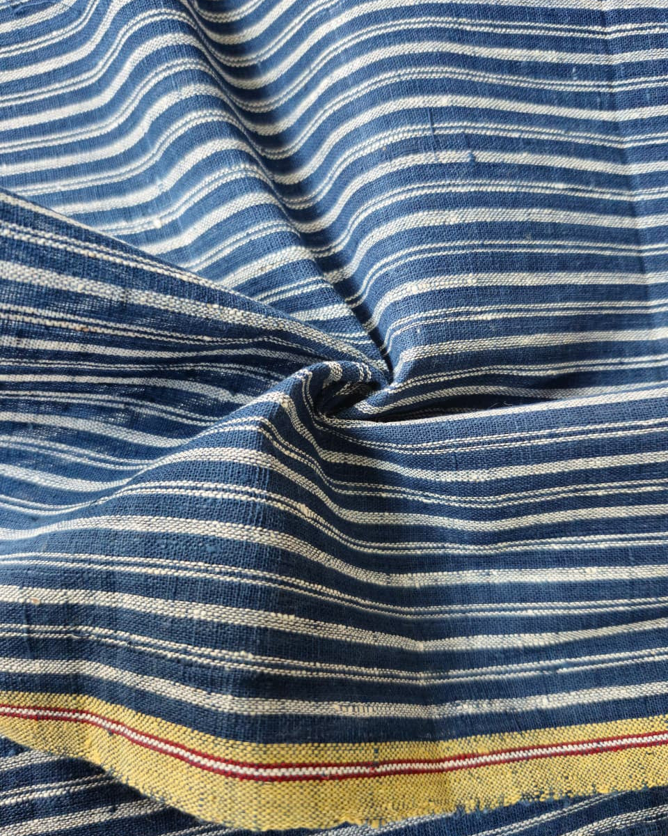 A close-up of a striped textile with alternating navy and off-white horizontal bands, gathered in the center. A yellow woven border with thin red and blue stripes is visible along the bottom edge.
