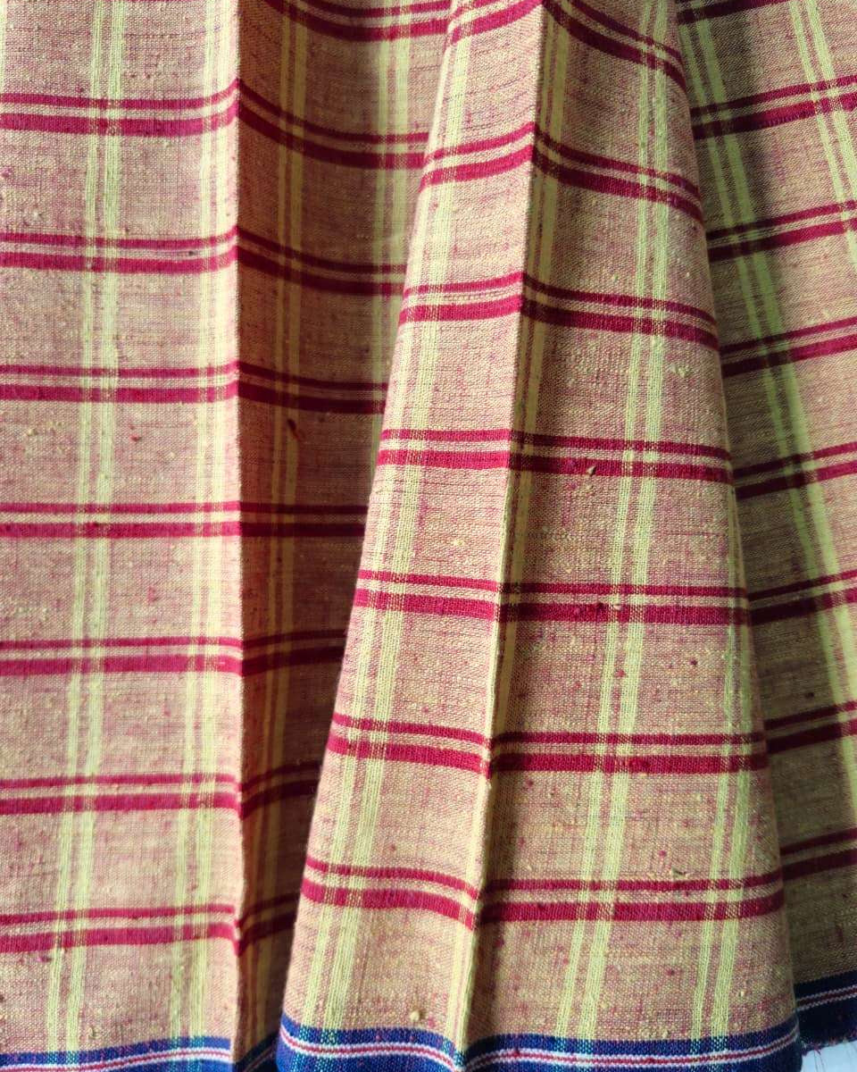 A close-up of a plaid fabric, likely a skirt or textile panel, featuring a beige base with red and light green stripes, finished with a blue and white striped border at the bottom.