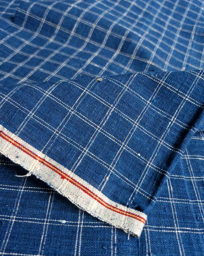 A close-up of blue fabric with a white windowpane plaid pattern and a folded hem showing a red and white striped inner edge.