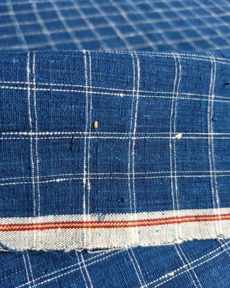 A close-up of blue woven fabric with a white grid plaid pattern and a frayed edge featuring a thin red stripe.