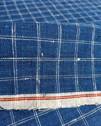 A close-up of blue woven fabric with a white grid plaid pattern and a frayed edge featuring a thin red stripe.