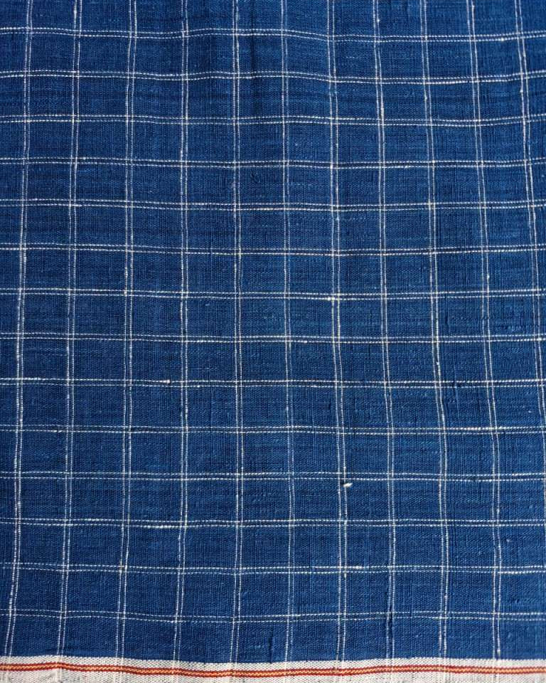 A close-up of a blue woven fabric with a white grid plaid pattern and a narrow orange-red stripe along the bottom edge.