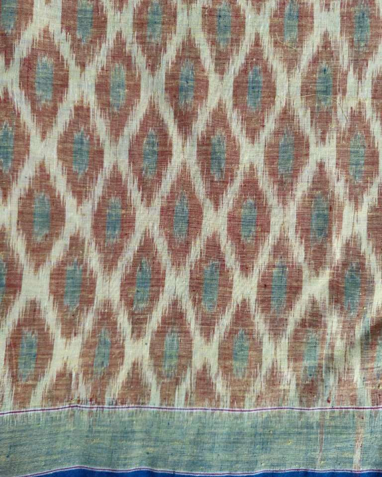 A close-up of a woven textile with an ikat-style diamond pattern in muted terracotta and pale blue on a warm beige ground, with a blue and green border along the bottom edge.
