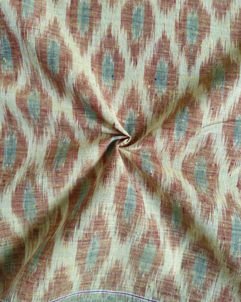 A close-up of patterned fabric with a dusty rose and beige ikat-style motif and muted blue-green accents, gathered in the center to show texture and drape.